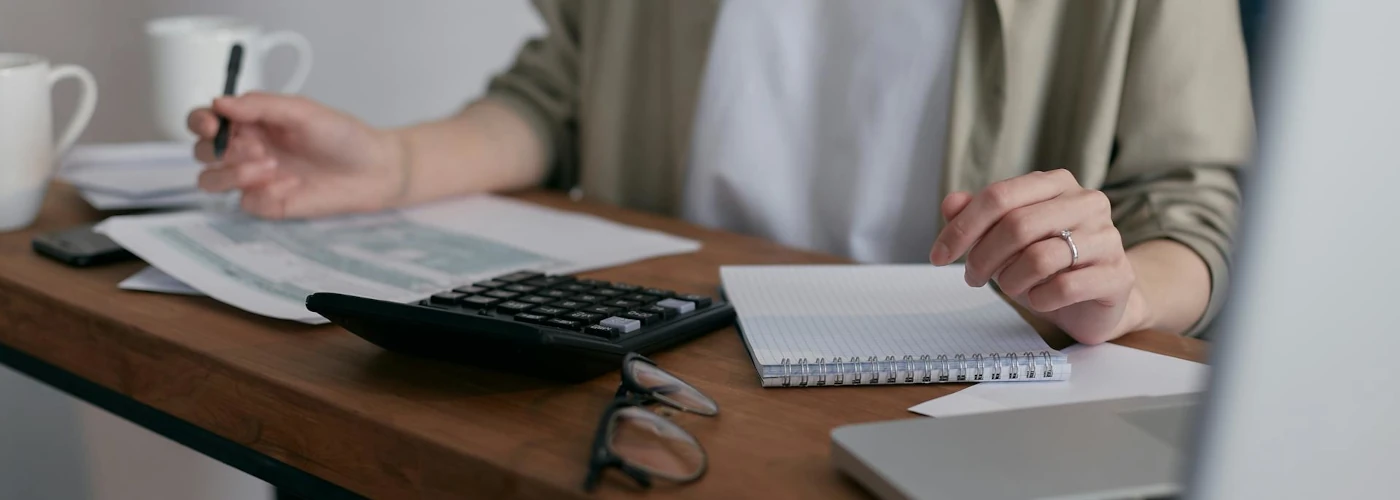 woman at desk holding a pen to paper with glasses, laptop and calculator.