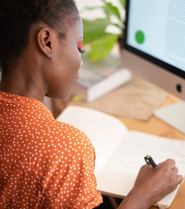 littlegreenbuttononscreen-portrait a black woman sitting at her desk writing in a notebook with a comptuter monitor in the background displaying the little green button panic alarm.